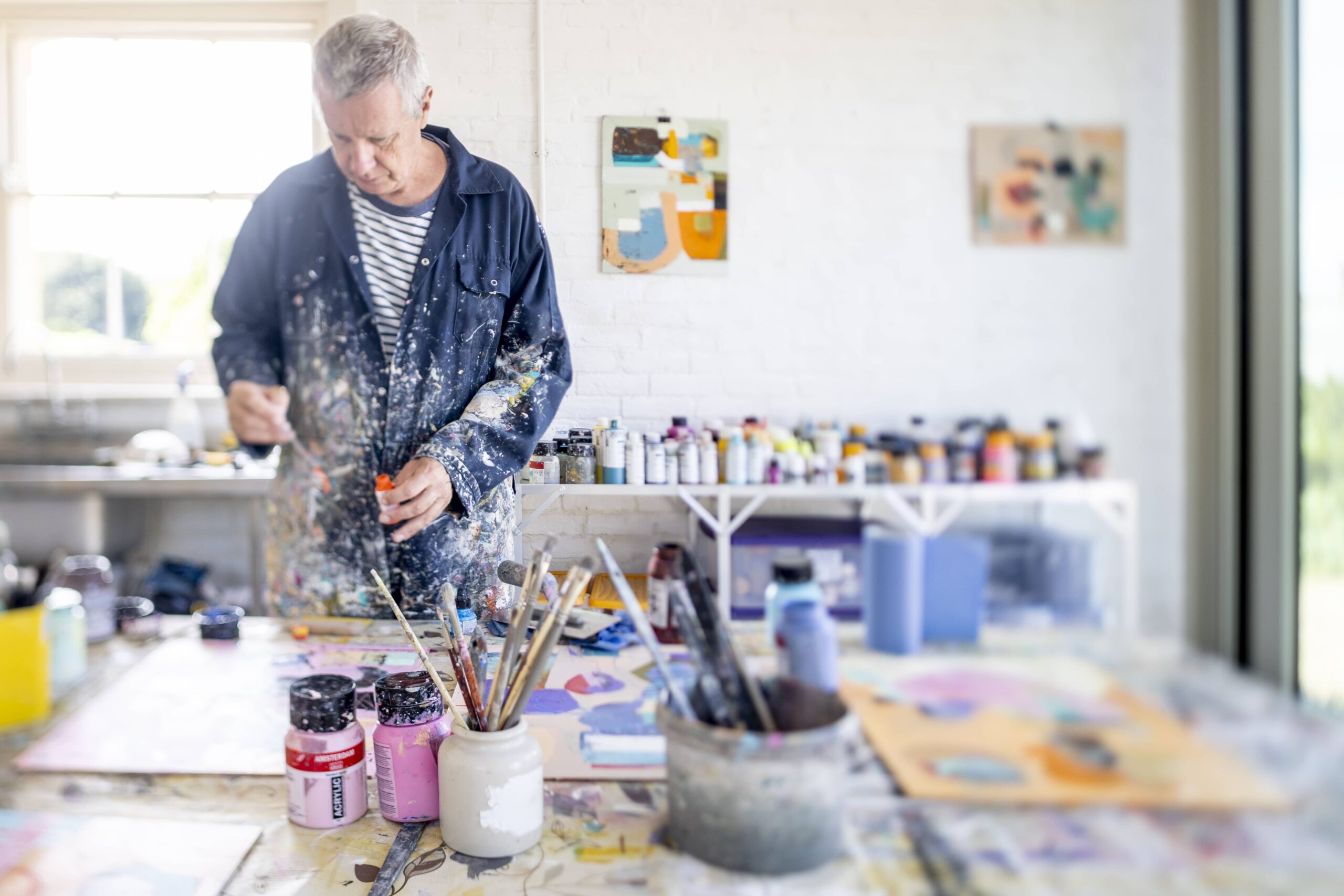 A man in painted overalls, surrounded by paint brushes, pots in a natural lit room