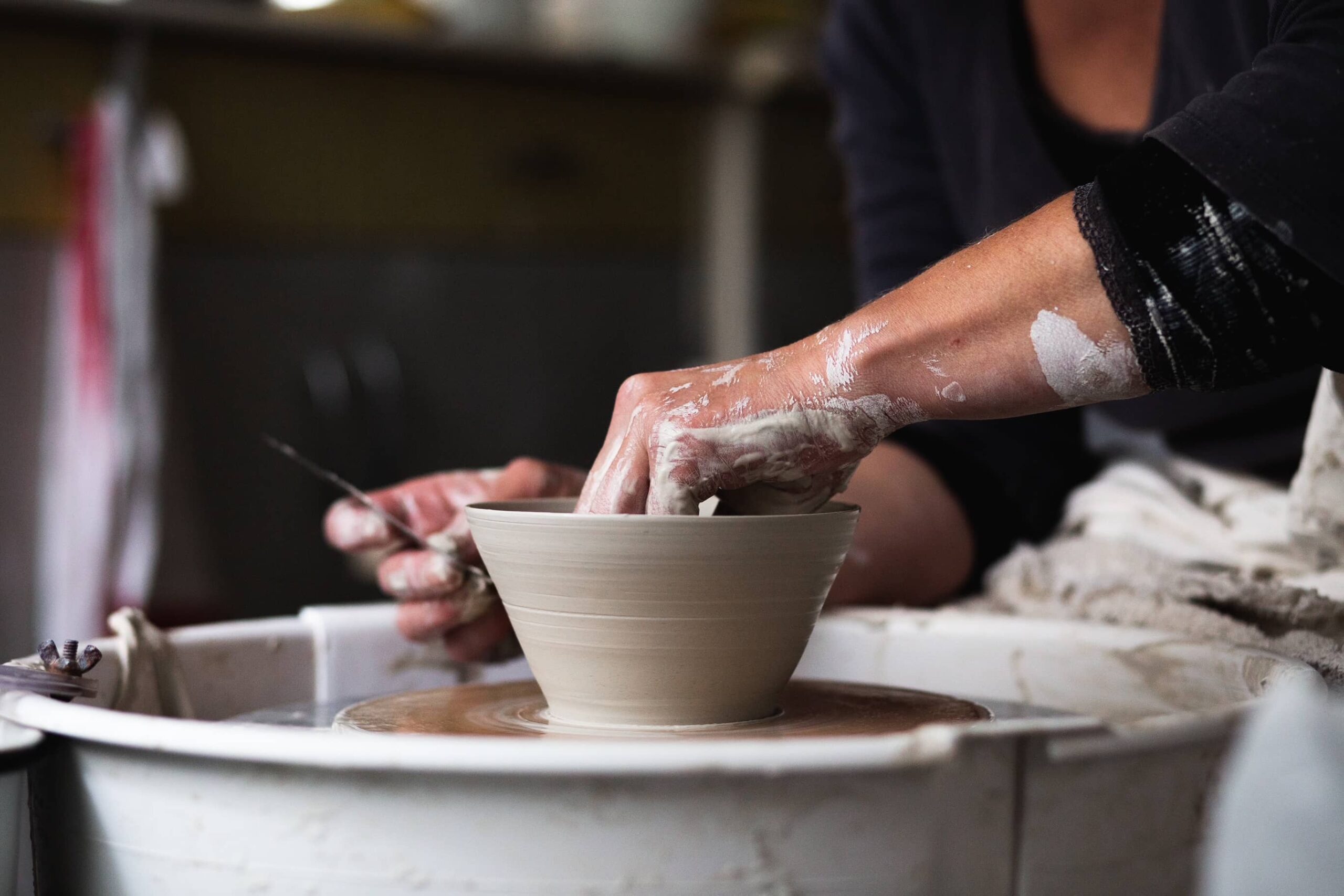 A photograph of a pottery wheel with a woman's hand making a small bowl from a grey clay. 