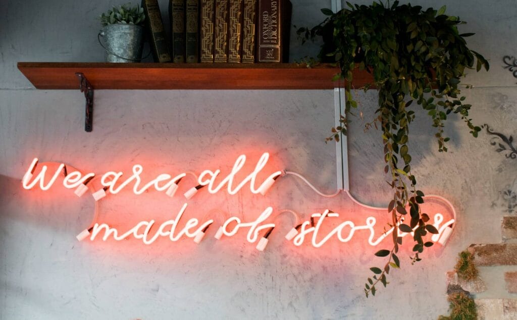 Neon sign saying we are all made of stories, on a wall under a bookshelf