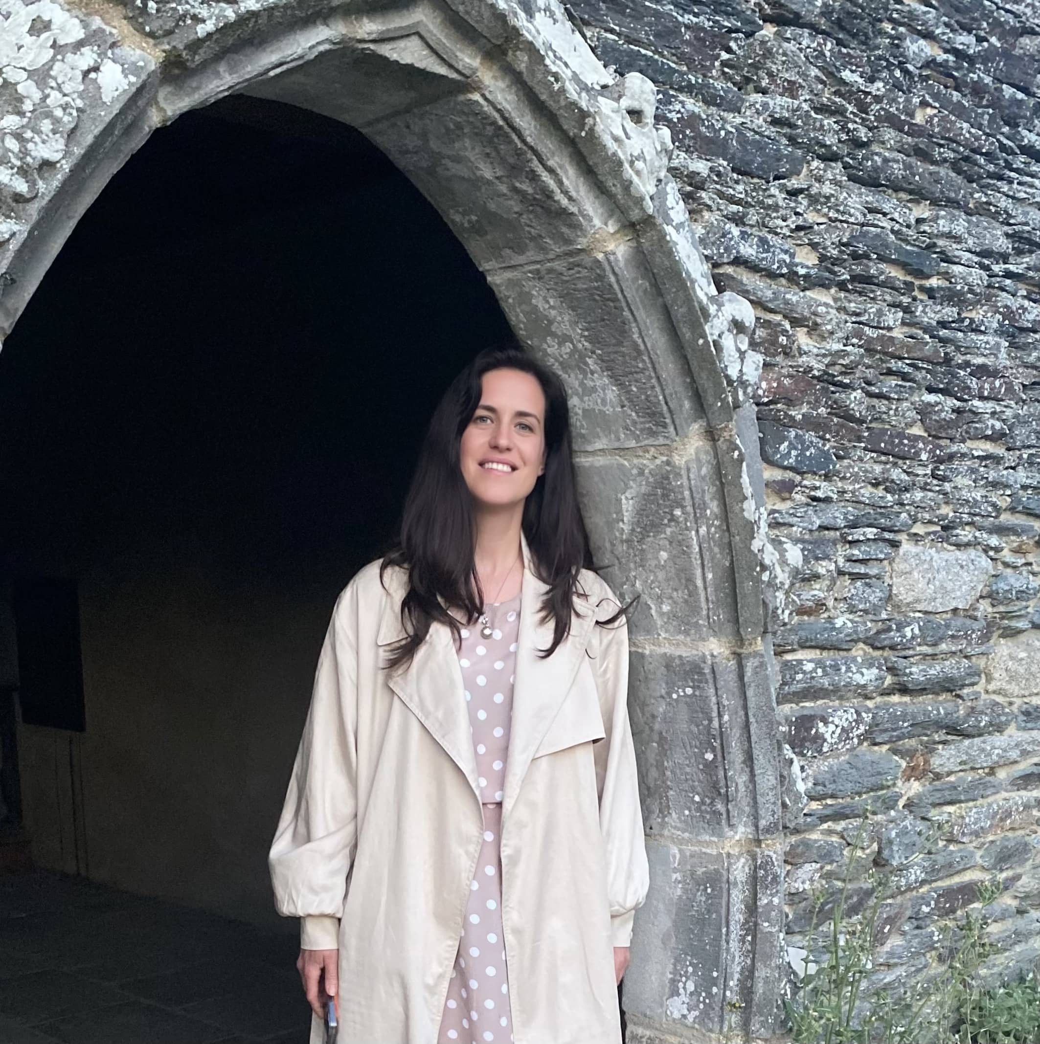 A picture of Jo. She wears a long white coat and is smiling in a stone archway.