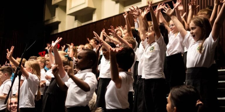 Image showing pupils singing with their hands in the air - with Southampton and IW Music hub logo overlaid