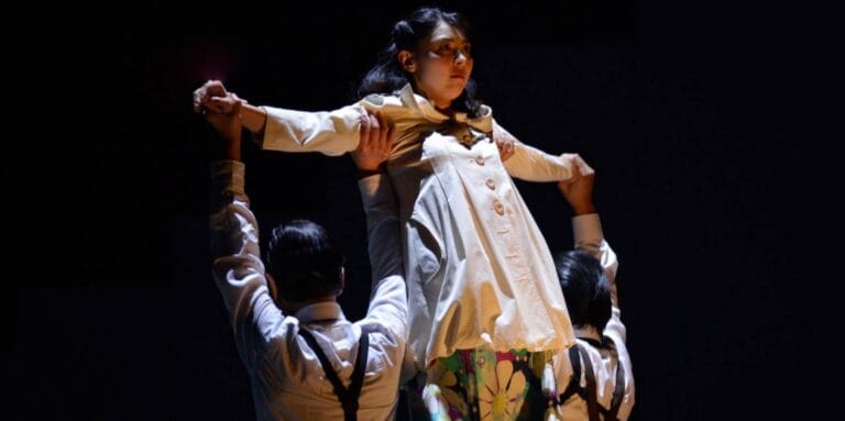 Two men on stage lifting a woman, with dark background and StoneCrabs logo overlaid on top