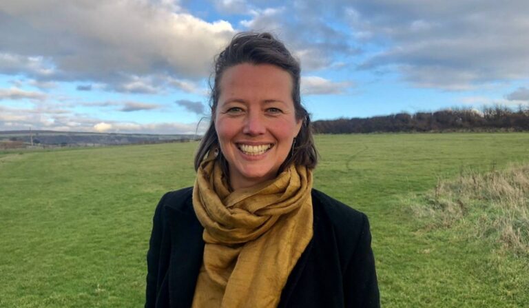 Charity Garnett is smiling as she's stood in a field with blue skies, wearing a yellow scarf and black cardigan.