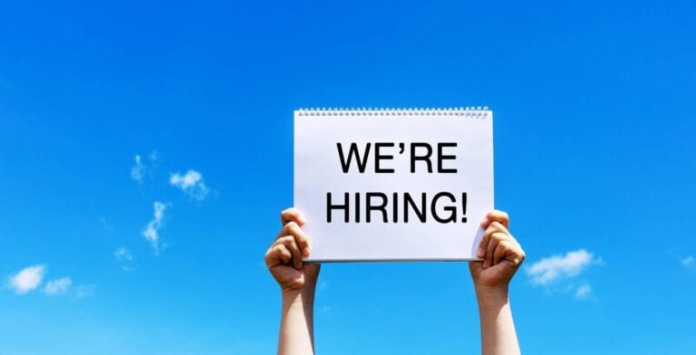Two hands holding up a sign saying 'We're Hiring' against a sky backgound