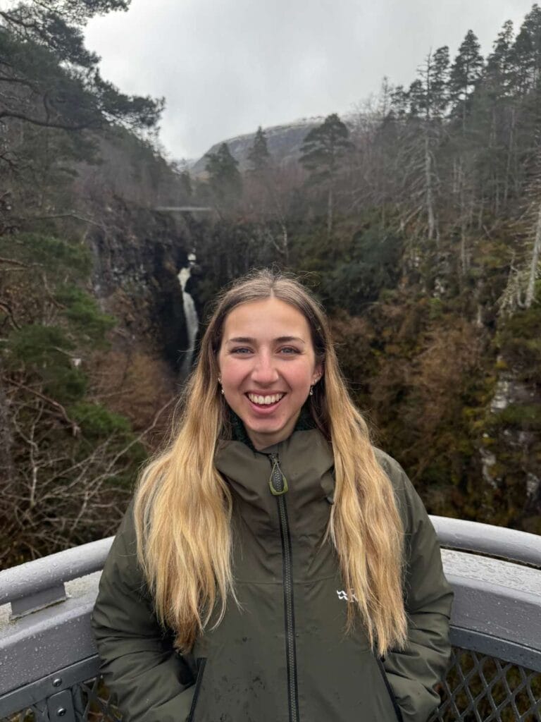 A photo of Amy. She's smiling as she stands in front of a mountain landscape somewhere in Scotland.
